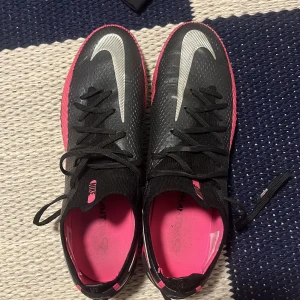 Svarta och rosa fotbollsskor från Nike - Snygga fotbollsskor från Nike i svart och rosa med silverdetaljer. Skorna har snörning och en slitstark sula med dobbar för bra grepp på planen. Perfekta för fotbollsträning och matcher. Skorna kostar 3000kr nypris och är 8,5 skick av 10 och är elite.