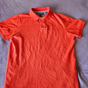 Orange pikétröja från Tommy Hilfiger - Säljer en snygg orange pikétröja från Tommy Hilfiger i storlek S. Tröjan har en klassisk krage med knappar och det ikoniska logotypmärket på bröstet. Perfekt för en stilren och avslappnad look.