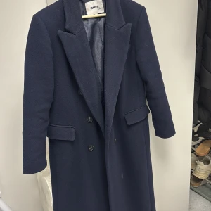 Mörkblå kappa från ONLY - Elegant mörkblå kappa från ONLY med klassisk design och knappar framtill. Perfekt för kyligare dagar med sin långa passform och stilrena look. Kappan har långa ärmar och är tillverkad i ett mjukt material som ger en bekväm känsla.