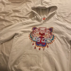Vit hoodie från Kenzo med tigerbroderi - Säljer en snygg vit hoodie från Kenzo med ett färgglatt tigerbroderi och texten 'Kenzo Paris' på framsidan. Hoodien har en klassisk passform med långa ärmar och en praktisk känguruficka. Perfekt för en stilren och bekväm look. Kan gå ner i pris vi snabba affärer