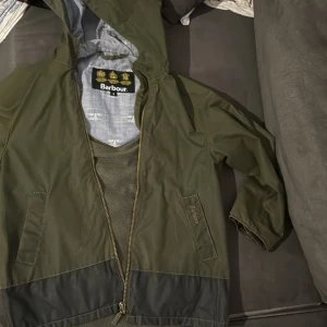 Grön jacka från Barbour Barn - Säljer en grön jacka från Barbour med luva och dragkedja. Jackan har ett mönstrat innerfoder och är perfekt för höstväder. Den har långa ärmar och en stilren design. Storleken är barn S. 