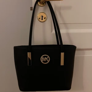 Svart handväska  -  Snygg svart handväska från Michael Kors med gulddetaljer och logotyp framtill. Väskan har två handtag och en dragkedja på baksidan för extra förvaring. Perfekt för att bära dina dagliga nödvändigheter med stil.❤️ inte säker på om den är äkta.