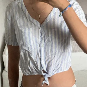Randig kortärmad blus - Säljer en stilren blus med blå och vita ränder. Blusen har korta ärmar och en knytning framtill som ger en trendig look. Perfekt för varma dagar!