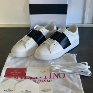 Valentino Garavani skor - Snygga vita sneakers från Valentino Garavani med en bred svart rem över snörningen. Skorna har en klassisk design med rund tå och är tillverkade i skinn. Perfekta för en stilren look, 