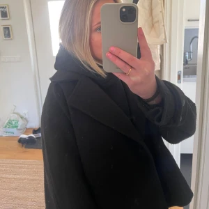 Svart kappa - Snygg svart kappa med klassisk krage och långa ärmar. Perfekt för kyligare dagar och men även bra inför våren och ger en stilren look. Passar bra till både vardag och mer uppklädda tillfällen.