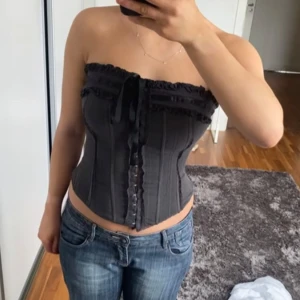 Svart korsett med snörning - Snygg svart korsett med snörning bak och dekorativa volanger framtill. Axelbandslös design som ger en elegant look. Perfekt för att skapa en stilren och chic outfit.