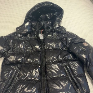 Moncler Maya  - Snygg svart glansig dunjacka från Moncler med huva och dragkedja. Jackan har ett klassiskt Moncler-märke på ärmen och en serietidningsetikett på insidan. Perfekt för kyliga dagar.