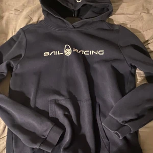 Mörkblå hoodie från Sail Racing - Säljer en snygg mörkblå hoodie från Sail Racing med vit logga på bröstet. Tröjan har en klassisk känguruficka och en bekväm huva. Perfekt för en avslappnad stil.