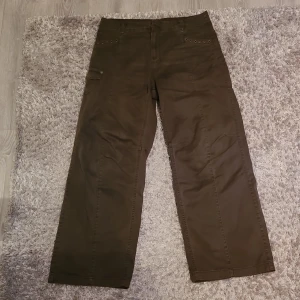 Bruna cargopants med detaljer - Snygga bruna cargopants med breda ben och coola nitar vid fickorna. Byxorna har en praktisk ficka med lock på sidan och är perfekta för en avslappnad stil. De är tillverkade i ett slitstarkt material och har en midwaist passform.