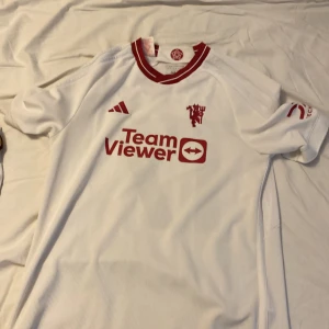 Vit fotbollströja med röd text - Säljer en vit Manchester United tröja. Tröjan har korta ärmar och ett tryck med namnet 'Garnacho' och nummer 17 på baksidan. Perfekt för fotbollsfans!