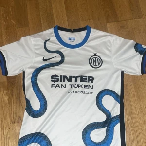 Vit och blå Inter Milano fotbollströja - Säljer en vit och blå Inter Milano fotbollströja från Nike. Tröjan har ett unikt ormmönster i blått och svart, med klubbens emblem och sponsorloggor på framsidan. Den har korta ärmar med blå detaljer vid ärmsluten.
