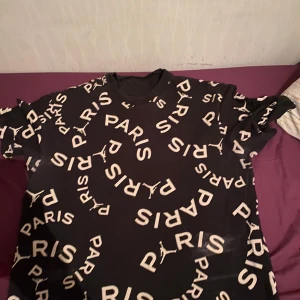 Svart t-shirt med Paris-tryck från Jordan - Snygg svart t-shirt från Jordan med stort Paris-tryck över hela tyget. Perfekt för dig som gillar sportig stil och vill ha något unikt i garderoben. T-shirten har korta ärmar och en normal passform.