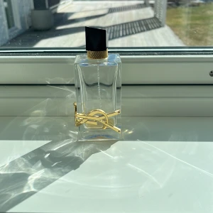 Parfymflaska från Yves Saint Laurent - Elegant parfymflaska från Yves Saint Laurent med en stilren, rektangulär design. Flaskan är genomskinlig med en svart kork och har en dekorativ guldfärgad logotyp på framsidan. Perfekt för den som söker en lyxig doftupplevelse.