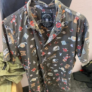 Mönstrad skjorta från Fabric Goods - Säljer en cool skjorta från Fabric Goods med ett unikt mönster av rymdmotiv och blommor. Skjortan har korta ärmar och knappar framtill. Perfekt för dig som vill sticka ut med en färgglad och lekfull stil.