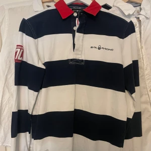 Randig pikétröja från Sail Racing - Säljer en stilren randig pikétröja från Sail Racing i marinblått och vitt med röd krage. Tröjan har långa ärmar och knappar framtill. Perfekt för en sportig look!