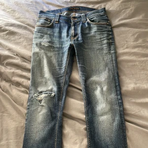 Blå jeans från Nudie Jeans - Snygga blå jeans från Nudie Jeans med slitna detaljer och en klassisk femficksdesign. Jeansen har en dragkedja och knappstängning samt en cool distressed look. Perfekta för en avslappnad stil. Hålen är ihopsydda med lapp bakom så man inte ser benen samt uppsydda till ungefär 32 i längd 