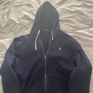Mörkblå zip hoodie från Polo Ralph Lauren - Säljer en snygg mörkblå zip hoodie från Polo Ralph Lauren med dragkedja och huva. Den har en broderad logga på bröstet och snören vid huvan. Perfekt för en avslappnad stil.