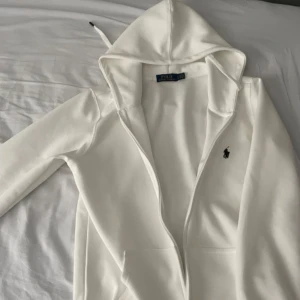 Vit hoodie från Ralph Lauren - Säljer en stilren vit hoodie från Ralph Lauren med dragkedja och det klassiska broderade logotypen på bröstet. Perfekt för en avslappnad look. Den har en justerbar huva och fickor framtill.