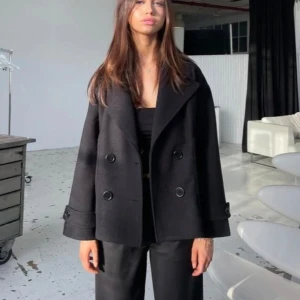 Svart kappa med dubbelknäppning - Elegant svart kappa med dubbelknäppning och breda slag. Jackan har långa ärmar med dekorativa knappar vid ärmsluten. Perfekt för en stilren look. Från design by si, köptes för 813💕