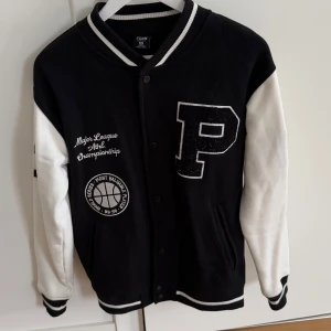 Svart och vit bomberjacka från FSBN - Säljer en svart och vit bomberjacka från FSBN med ett stort 'P' på bröstet och texten 'Major League Alpha Championship'. Jackan har knappar framtill och ribbade muddar i svart och vitt. Perfekt för en sportig look! Stor i storlek, mer som M/L