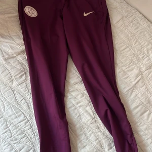 Lila träningsbyxor från Nike - Snygga lila träningsbyxor från Nike med elastisk midja och logotyp på benet. Perfekta för sport och fritid. Byxorna har en smal passform och är tillverkade i ett bekvämt syntetmaterial.