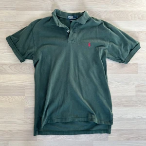 Grön pikétröja från Ralph Lauren - Säljer en klassisk grön pikétröja från Ralph Lauren med korta ärmar och den ikoniska broderade loggan i rött på bröstet. Tillverkad i 100% bomull för en bekväm passform. Perfekt för en stilren look!