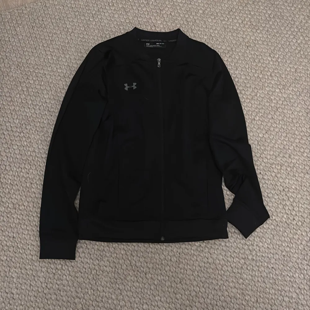 Säljer en stilren svart träningströja från Under Armour. Jackan har en dragkedja framtill och är långärmad. Perfekt för en sportig look eller avslappnade dagar.. Takit.