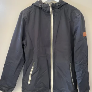 Mörkblå jacka från Lyle & Scott - Snygg mörkblå jacka från Lyle & Scott med dragkedja och huva. Jackan har en diskret logga på ärmen och praktiska fickor med dragkedja. Perfekt för en stilren look. Hör av dig om du vill veta mer. Pris kan diskuteras vid snabb affär! 