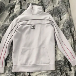 Vit tröja med hög krage och broderad logga - Säljer en stilren vit tröja med hög krage och broderad logga på bröstet. Tröjan har svarta och röda linjer längs ärmarna och en praktisk magficka framtill. Perfekt för en avslappnad stil.