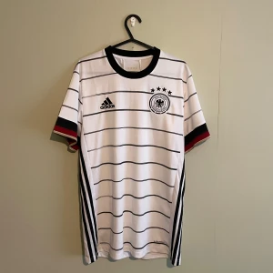 Tyskland fotbollströja från Adidas - Säljer en vit fotbollströja från Adidas med svarta och röda detaljer. Tröjan har korta ärmar och ett randigt mönster. Den har även ett tryck med texten 'Deutscher Fussball-Bund' och en svart örn på bröstet. Perfekt för fotbollsfans!