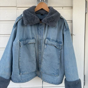 Blå jeansjacka med päls  - Säljer en snygg blå jeansjacka med grå fuskpäls på krage och ärmslut. Jackan har en dragkedja framtill och stora fickor med lock. Perfekt vår jacka ! vet inte storlek men passar mig som är S-M