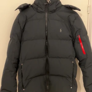 Svart dunjacka från Polo Ralph Lauren - Säljer en snygg svart dunjacka från Polo Ralph Lauren. Jackan har en dragkedja framtill och en praktisk huva. Den har en ficka med dragkedja på ärmen och man kan även ta bort luvan