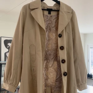 Beige trenchcoat med mönstrat foder - Snygg beige trenchcoat med stora knappar och ett unikt mönstrat foder. Jackan har långa ärmar och en klassisk krage. Perfekt för vår och höst. Mkt fint skick 