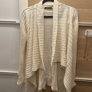 Créme vit ribbad kofta från Gina Tricot - Säljer en stilren beige kofta från Gina Tricot. Den är ribbad och har en öppen design som ger en avslappnad look. Perfekt för lager-på-lager under kyligare dagar.