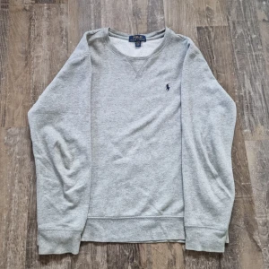 Grå sweatshirt från Polo Ralph Lauren - Säljer en klassisk grå sweatshirt från Polo Ralph Lauren med det ikoniska logotypbroderiet på bröstet. Tröjan har långa ärmar och en rund halsringning. Perfekt för en avslappnad stil.