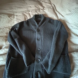 Grå stickad cardigan med knappar - Säljer en stilren grå stickad cardigan med stora knappar framtill från merley Stockholm . Cardiganen har en ribbad struktur och långa ärmar, perfekt för kyliga dagar. Den har också praktiska fickor på sidorna.