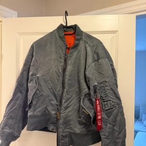 Grå/blå bomberjacka från Alpha Industries - Säljer en klassisk grå bomberjacka från Alpha Industries med orange foder. Jackan har dragkedja framtill och ribbade muddar vid ärmslut och nederkant. Den har en ikonisk röd tagg på ärmen och flera praktiska fickor.  Finns fläckar  passar både S/M/L
