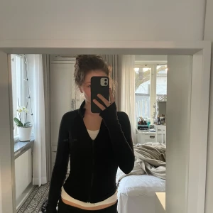 Trendiga Gina tröjan - Säljer en stilren svart hoodie med dragkedja. Den har långa ärmar och en åtsittande passform som framhäver figuren. Den är endast använd ett fåtal gånger och saknar defekter