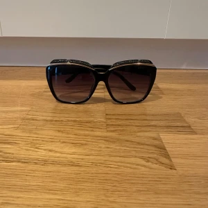 Svarta solglasögon från Dior - Snygga svarta solglasögon från Dior med en elegant fyrkantig form. De har mörka glas och en subtil glittrig detalj på bågarna. Perfekta för att ge en lyxig touch till din stil. Passar främst tjejer.