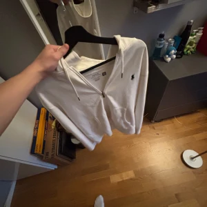Vit hoodie från Polo Ralph Lauren - Säljer en stilren vit hoodie från Polo Ralph Lauren med dragkedja och klassisk logga på bröstet. Perfekt för en avslappnad look. Den har en justerbar huva och praktiska fickor framtill. Passar bra till både jeans och joggers.
