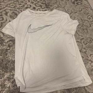 Vit t-shirt från Nike - Säljer en vit t-shirt från Nike med en stilren logga på framsidan. Perfekt för träning eller en avslappnad dag. T-shirten har korta ärmar och är gjord i ett mjukt material.