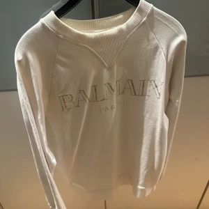 Vit tröja från Balmain - Säljer en stilren vit tröja från Balmain med deras logga tryckt på framsidan. Tröjan har långa ärmar och en ribbad krage som ger en klassisk look. Perfekt för dig som vill ha något enkelt men ändå exklusivt.