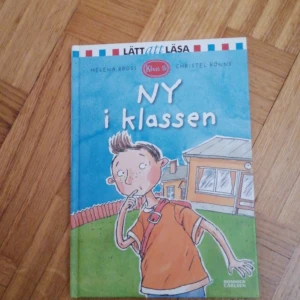 Ny i klassen - Boken 'Ny i klassen' av Helena Bross och Christel Rönns är en del av serien 'Lätt att läsa'. Den handlar om en ny elev i klassen och är perfekt för nybörjarläsare. Omslaget visar en tecknad pojke framför en skola.