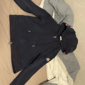 Mörkblå hoodie från Moncler - Säljer en mörkblå hoodie från Moncler med dragkedja och huva. Jackan har Moncler-logga på ärmen och ribbade muddar. Perfekt för dig som vill ha en stilren och sportig look.