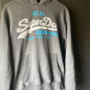 Grå hoodie från Superdry - Säljer en grå hoodie från Superdry med tryck i vitt och blått på framsidan. Tröjan har en klassisk känguruficka och en bekväm huva med dragsko. Perfekt för en avslappnad stil.