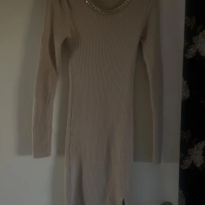 Beige ribbad klänning med kedjedetalj - Snygg beige ribbad klänning med lång ärm och en stilren kedjedetalj vid halsen. Perfekt för en elegant look. Klänningen har en liten slits nedtill för extra stil. aldrig använt.