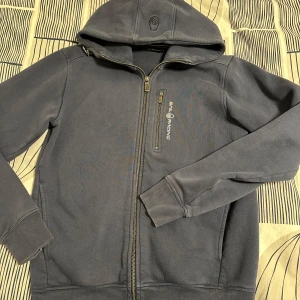 Mörkblå huvtröja från Sail Racing - Säljer en snygg mörkblå huvtröja från Sail Racing. Tröjan har en dragkedja framtill och en praktisk bröstficka med dragkedja. Perfekt för kyliga dagar med sin bekväma passform och stilrena design.