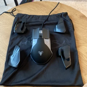 Svart gamingmus med utbytbara sidopaneler - Säljer en svart gamingmus med flera utbytbara sidopaneler för anpassad ergonomi. Musen har en sladd och en logotyp framtill. Perfekt för gamingentusiasten som vill ha flexibilitet och komfort.