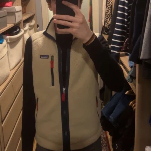 Patagonia - Säljer en beige fleeceväst från Patagonia med svarta detaljer och dragkedja. Västen har en klassisk passform och är perfekt för lager-på-lager under kyligare dagar. Den har en liten logga på bröstet och är både stilren och funktionell.