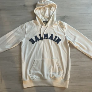 Vit hoodie från Balmain - Säljer en stilren vit hoodie från Balmain med blå text på bröstet. Tröjan har en klassisk känguruficka och justerbar huva . Perfekt för en avslappnad look.